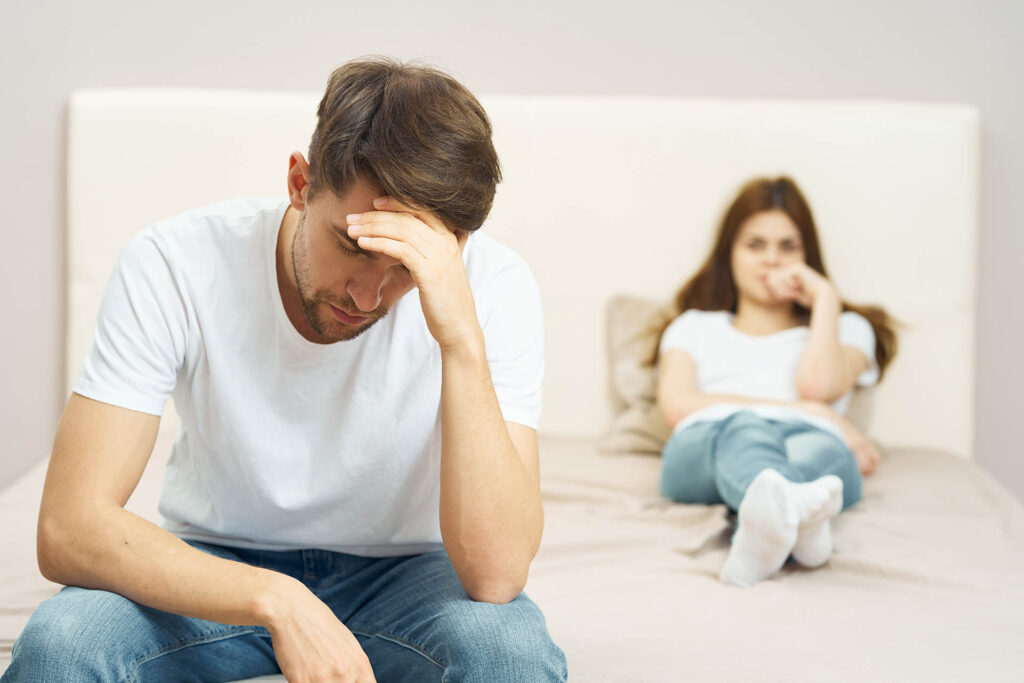 A man sitting on his bed looking frustrated with his partner sitting in the background.